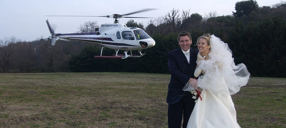 Couple de mariés souriant devant un hélicoptère de la société Nice Hélicoptères, robe de mariée et costume élégant, arrivée spectaculaire pour leur mariage, ambiance romantique et aventureuse en plein air.